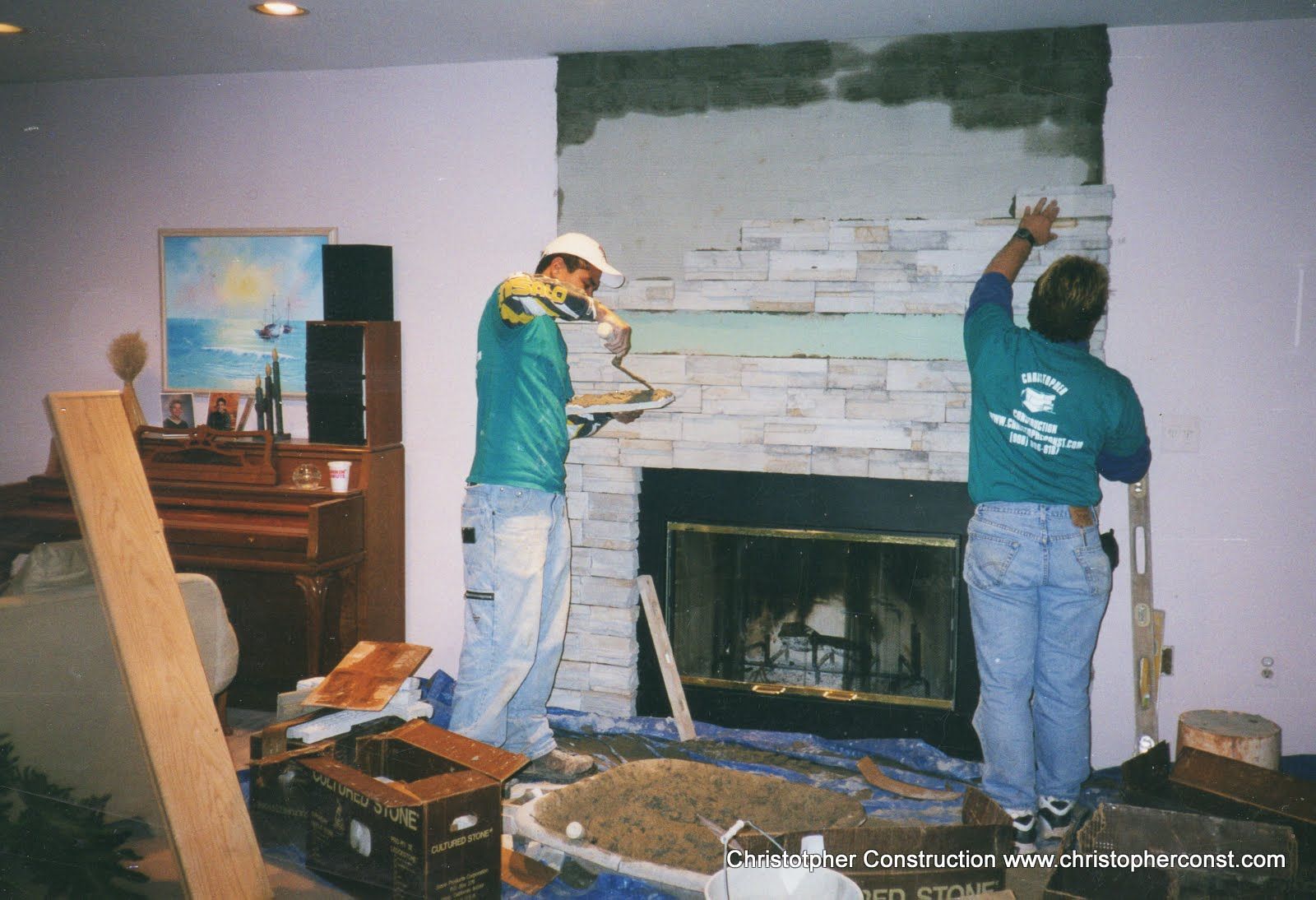 Two workers installing stone veneer around a fireplace in a room with a piano and tools scattered nearby