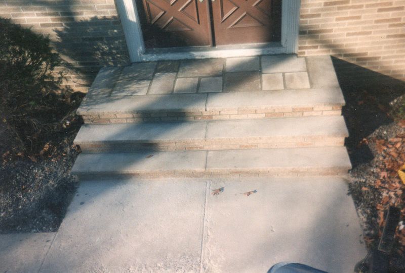 Concrete walkway leading to a front porch with two steps and a wooden door, bordered by mulch and shrubs