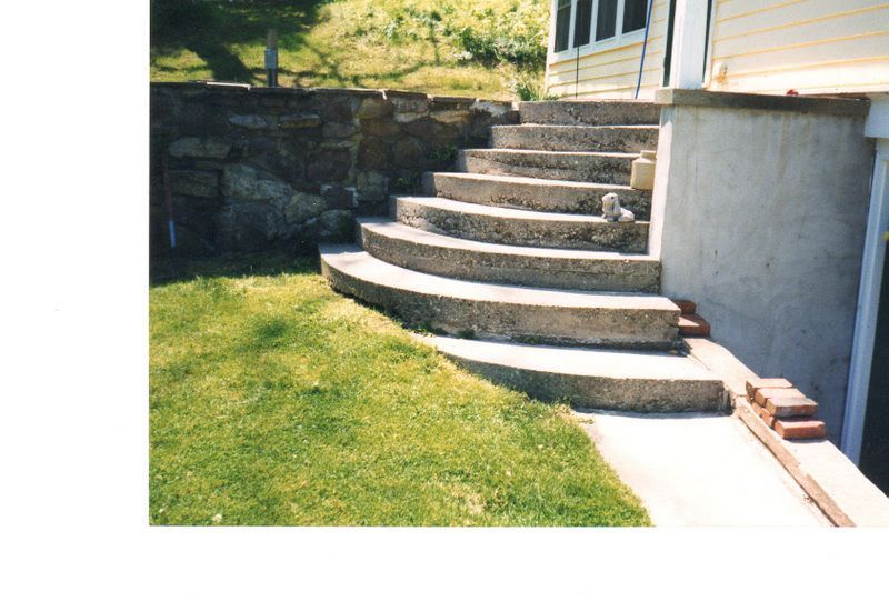 Curved stone steps beside a house, leading from a lawn to a raised patio with a retaining wall.
