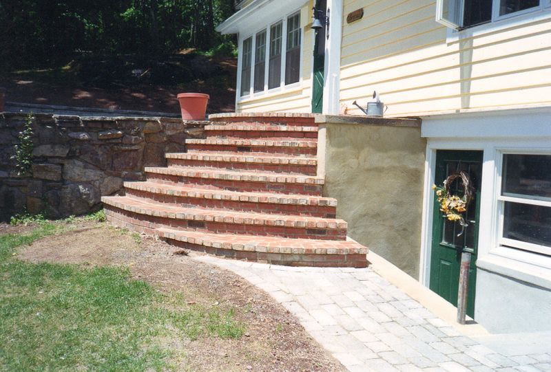 Brick steps leading to a house entrance beside a window and potted plant in a sunny yard