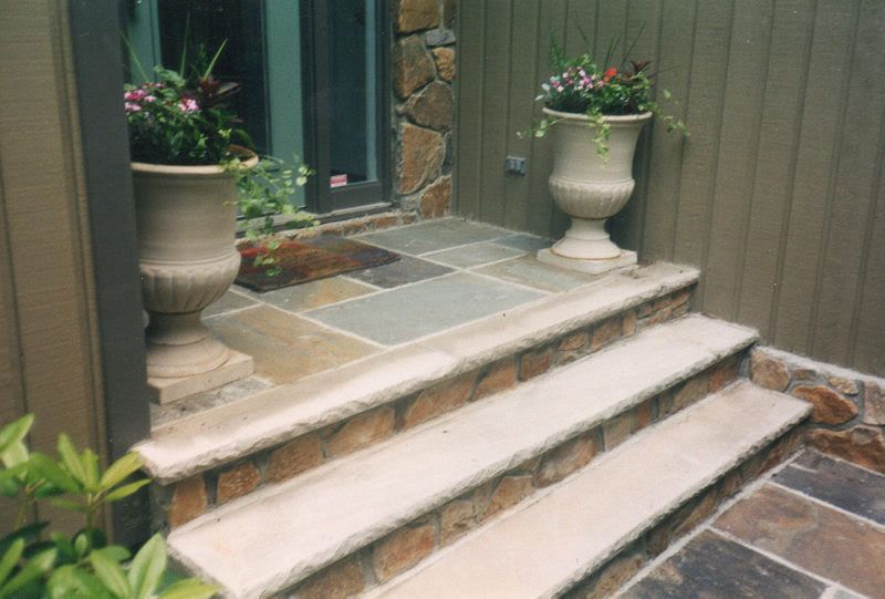 Front steps to a doorway with stone tiles and two large white planters on either side.