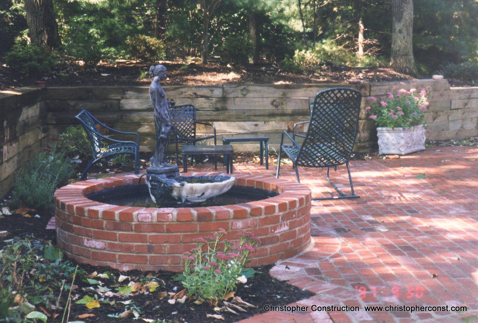 Brick circular fountain with black bird sculpture, surrounded by patio chairs and stone planters in a garden