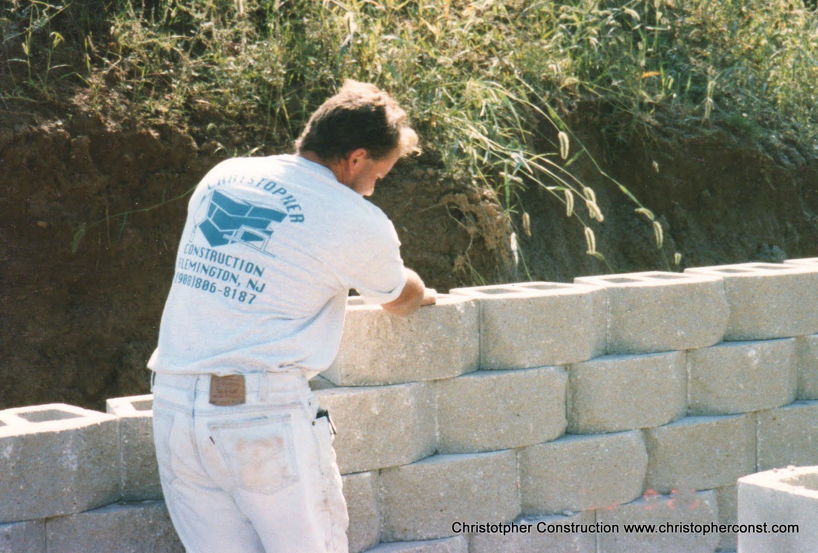 Man in white shirt and jeans stacking cinder blocks outdoors beside a dirt slope.
