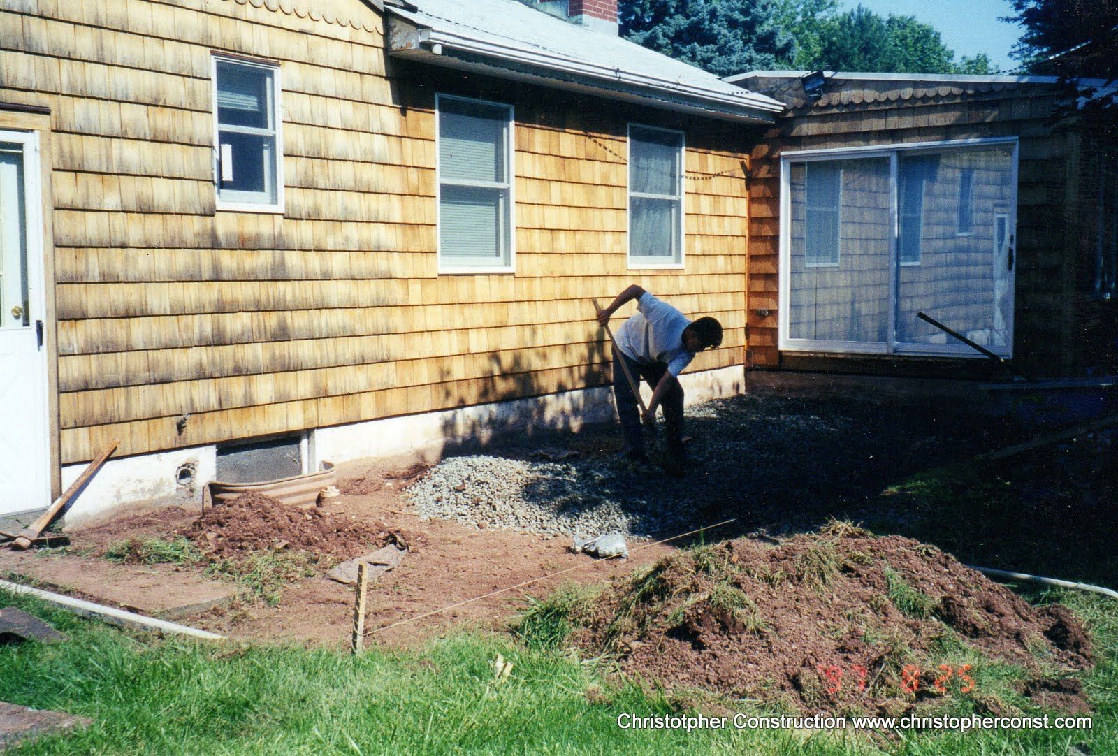Person working in a yard beside a yellow house, digging soil near the foundation.