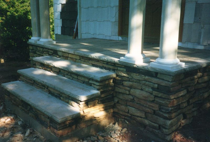 Front steps of a house with stone retaining wall, white columns, and sunlight on the porch