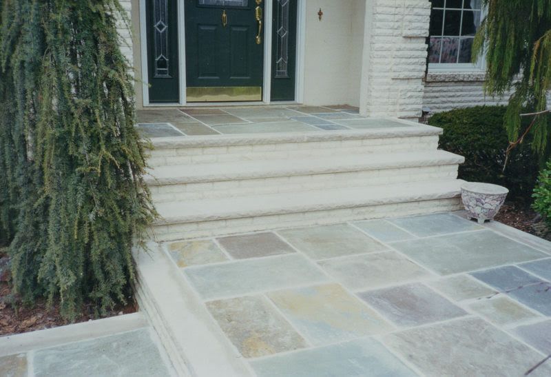 Stone front steps and walkway leading to a green front door, with a potted plant and hanging greenery.