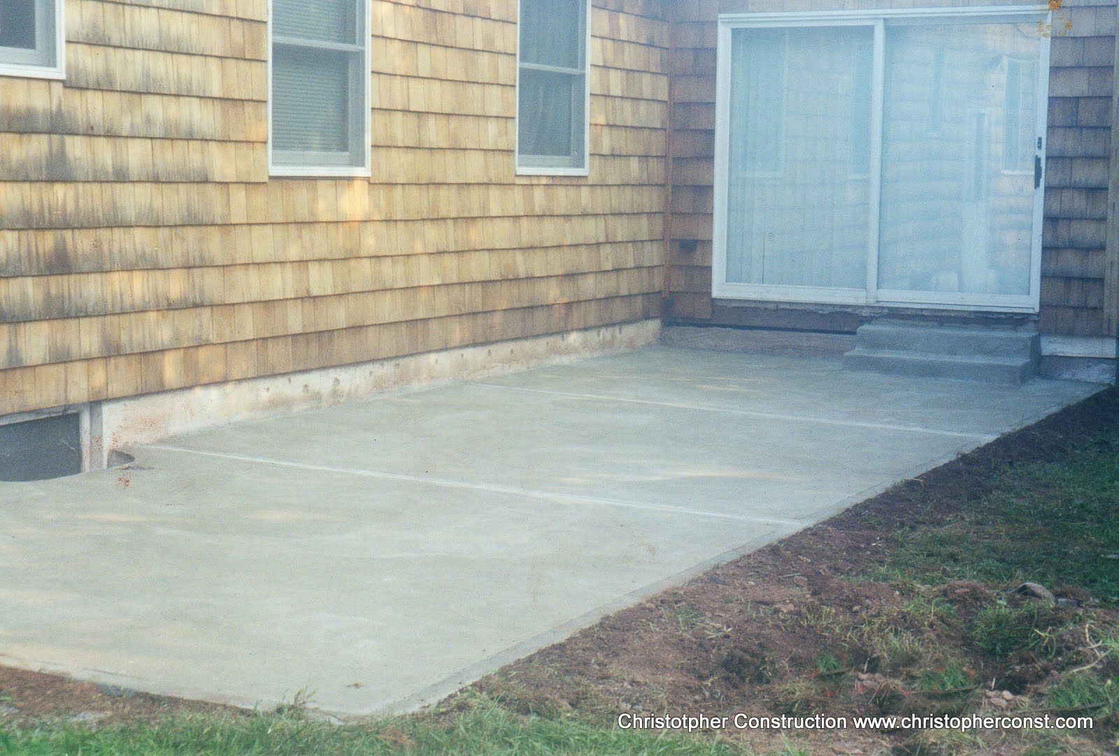 Concrete patio beside a tan shingled house with a white double door and small grass strip