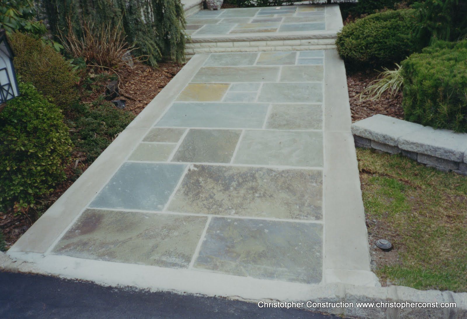 Stone walkway with large rectangular pavers bordered by concrete, leading through a landscaped garden
