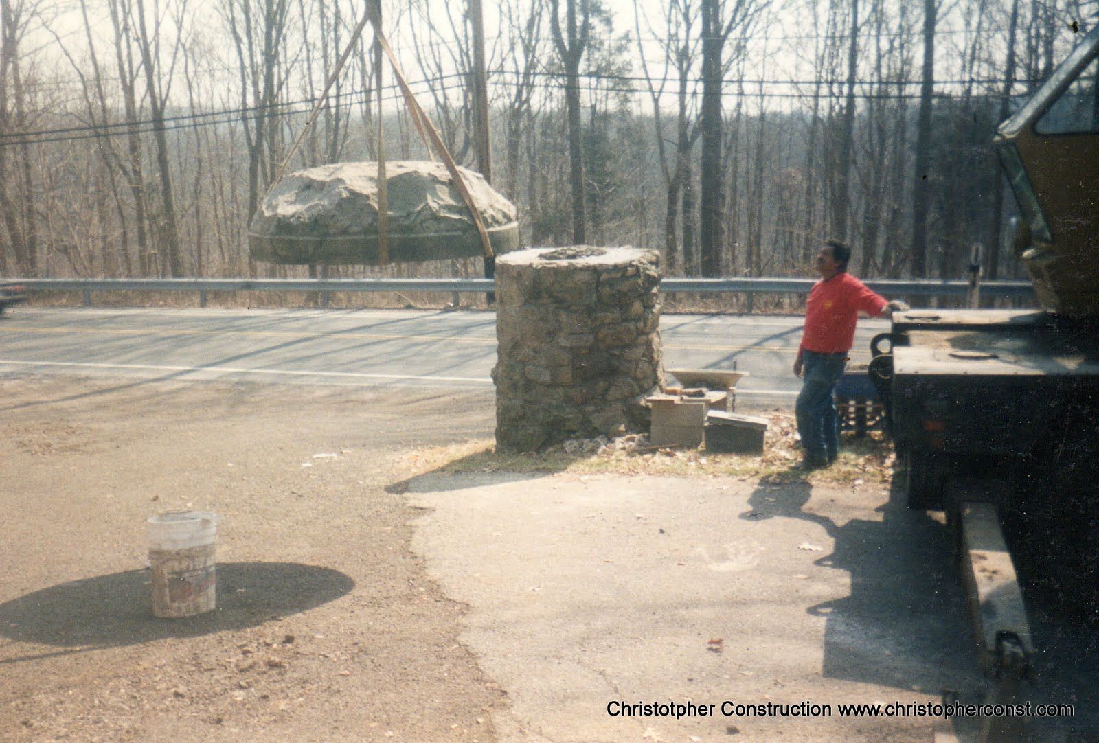 Person in a red jacket standing by a truck beside a stone pillar on a roadside in a wooded area