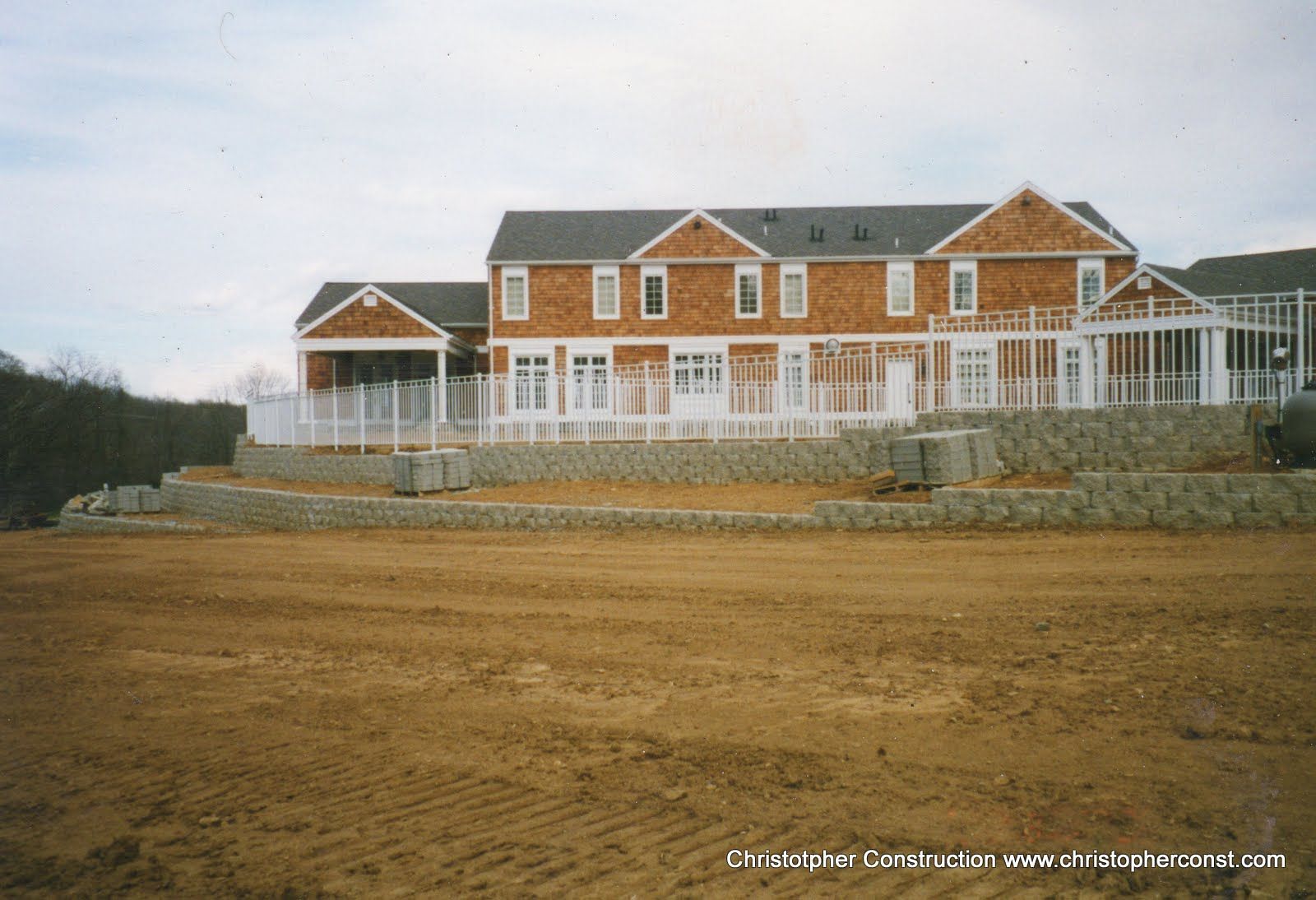 Large unfinished brick house with white trim and wraparound porch on a bare dirt lot under a cloudy sky