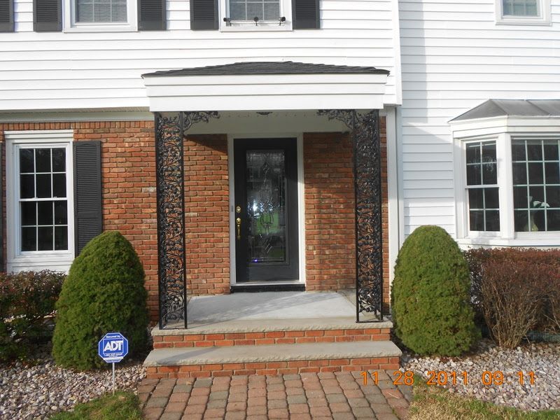 Front entrance of a brick and white house with a glass door, awning, and trimmed shrubs