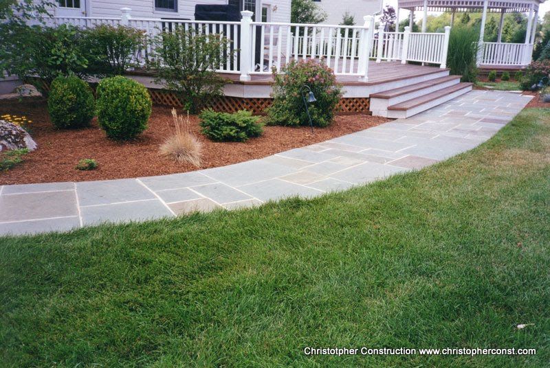 Curved concrete walkway leading to a white porch with steps, railing, shrubs, and green lawn