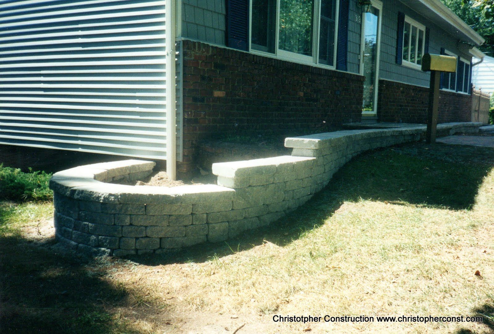 Curved stacked-stone retaining wall beside a house, with grass yard and brick steps leading up to the entry