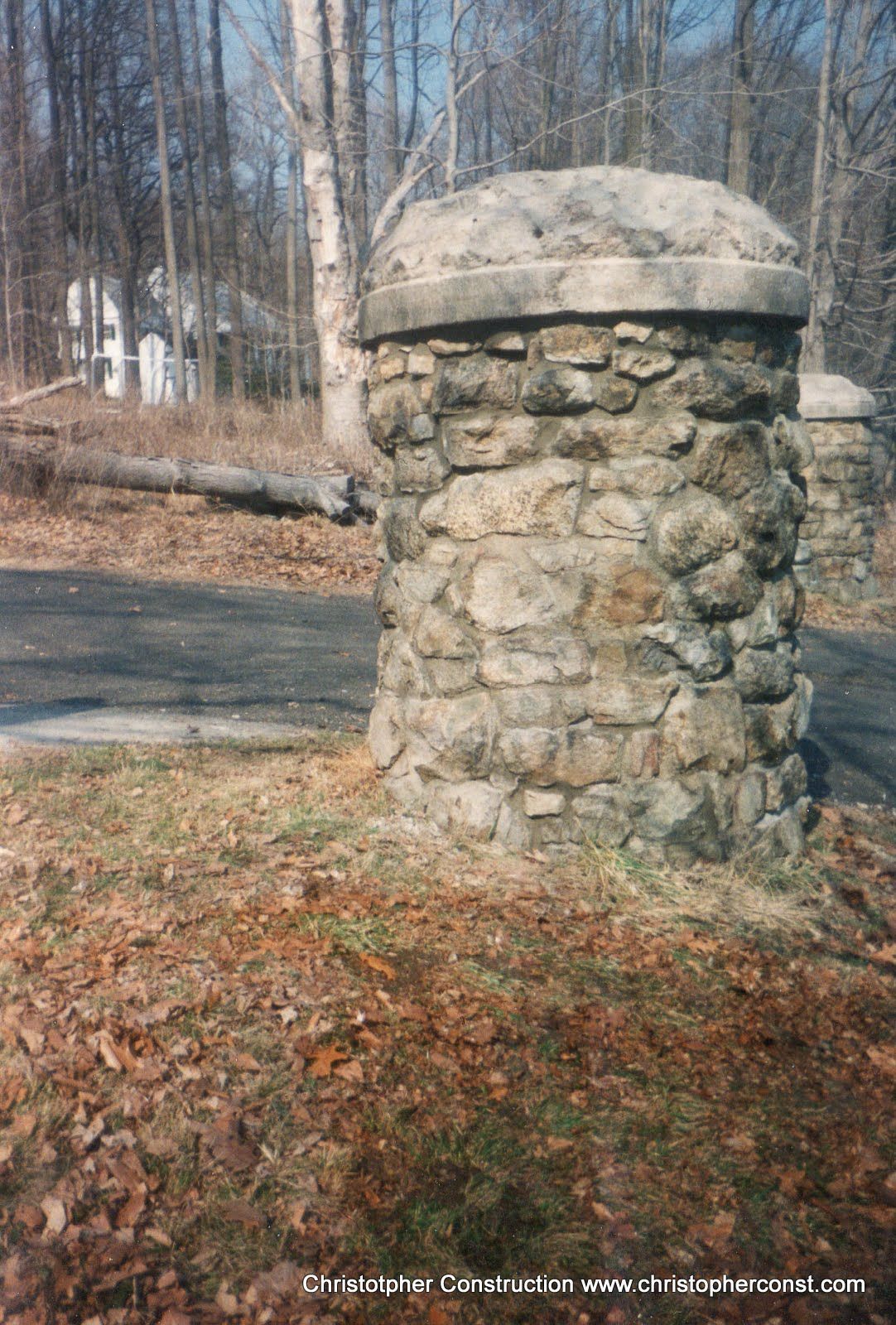 Stone pillar or monument in a wooded area beside a paved path, with fallen leaves on the ground.