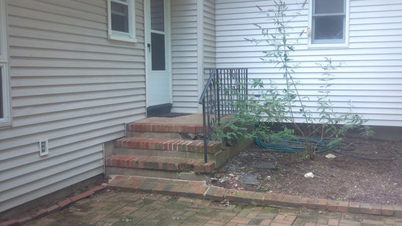 Brick steps and a small porch beside a beige house, leading to a white door with a railing and garden bed.
