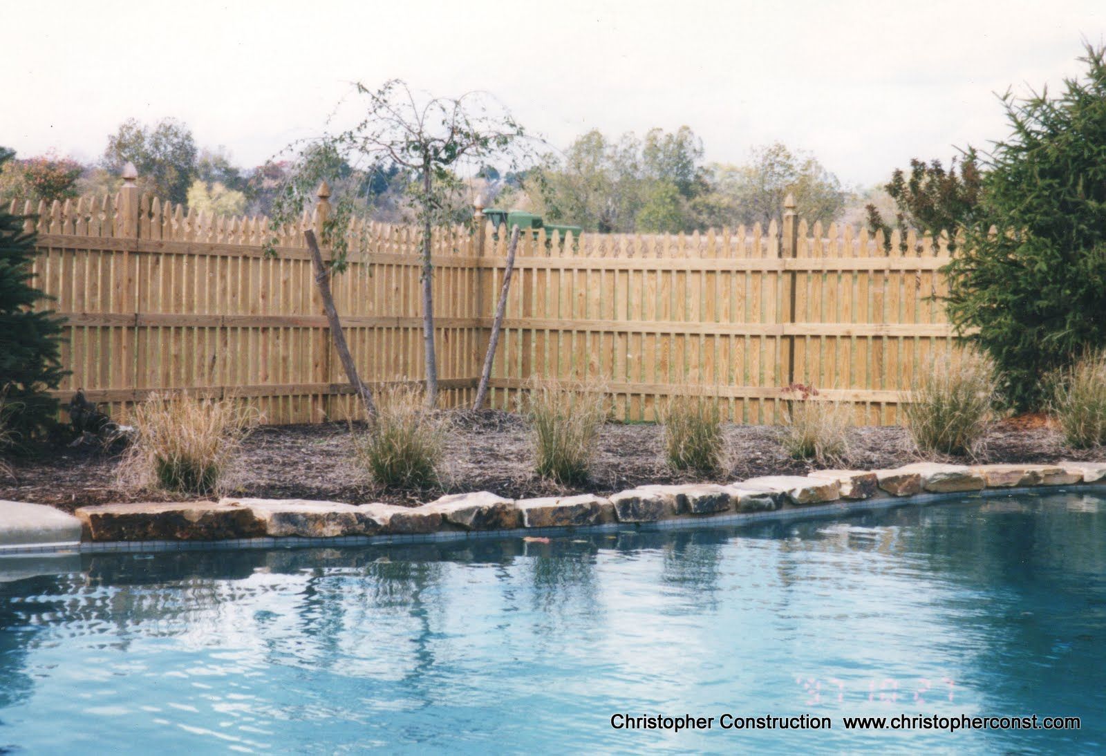 Backyard pool with blue water, rock border, and tall wooden fence with trees behind it