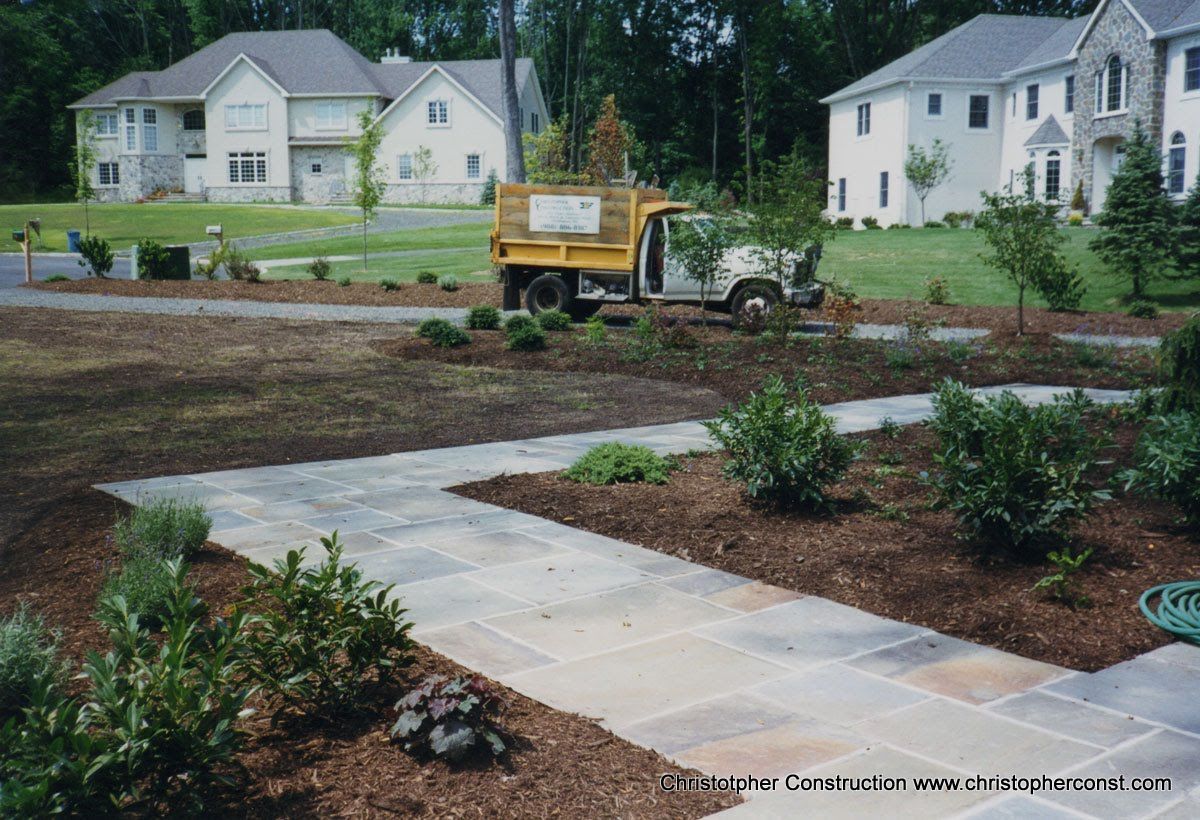 New residential landscaping with a yellow tractor, paved path, shrubs, and houses in the background