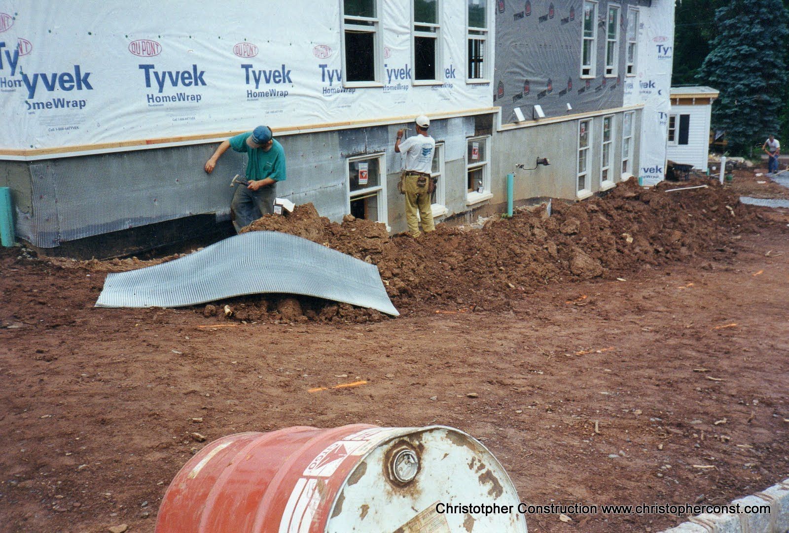 Construction site with workers, house under renovation, dirt piles, and a bent metal sheet in the foreground