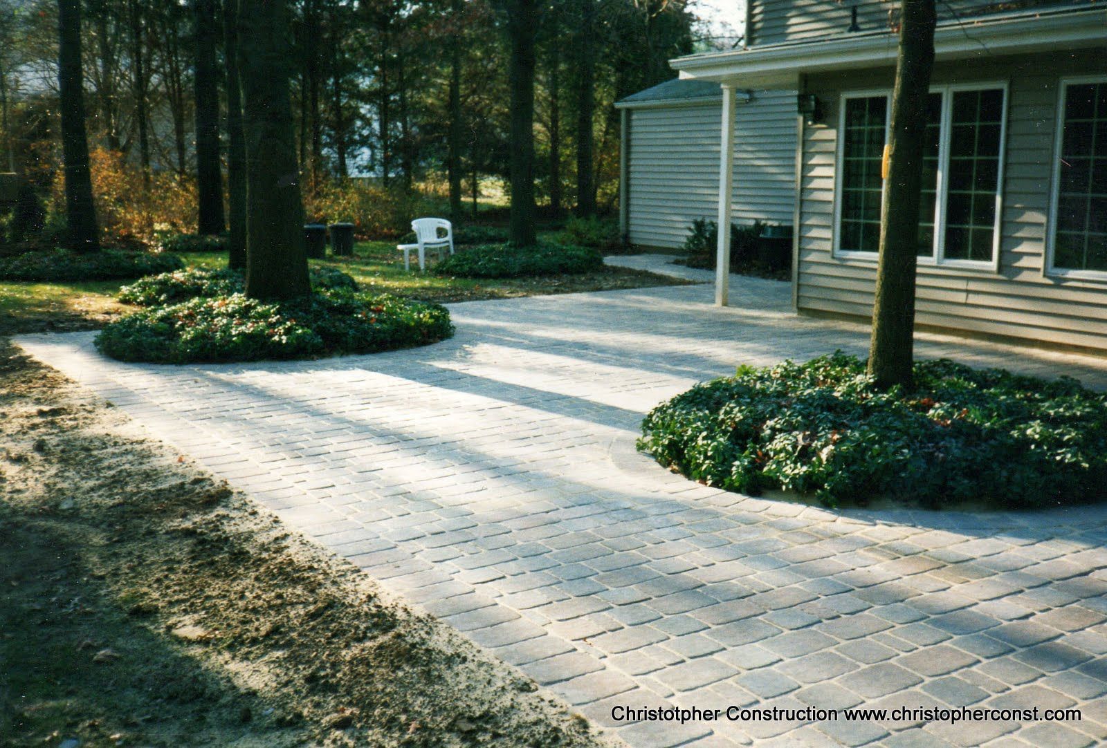 Stone patio and walkway beside a house, bordered by trees and flower beds in a wooded yard