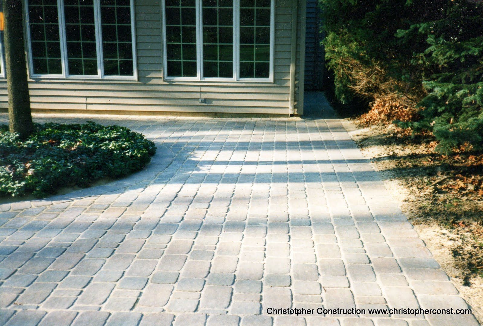 Paved brick walkway leading to glass doors, bordered by low shrubs and sunlit grass