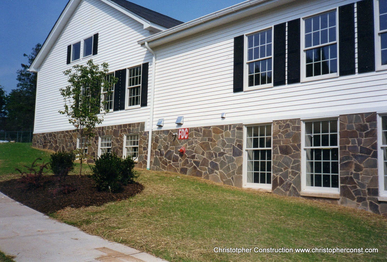 Suburban house exterior with white siding, stone foundation, black shutters, and a small front tree.