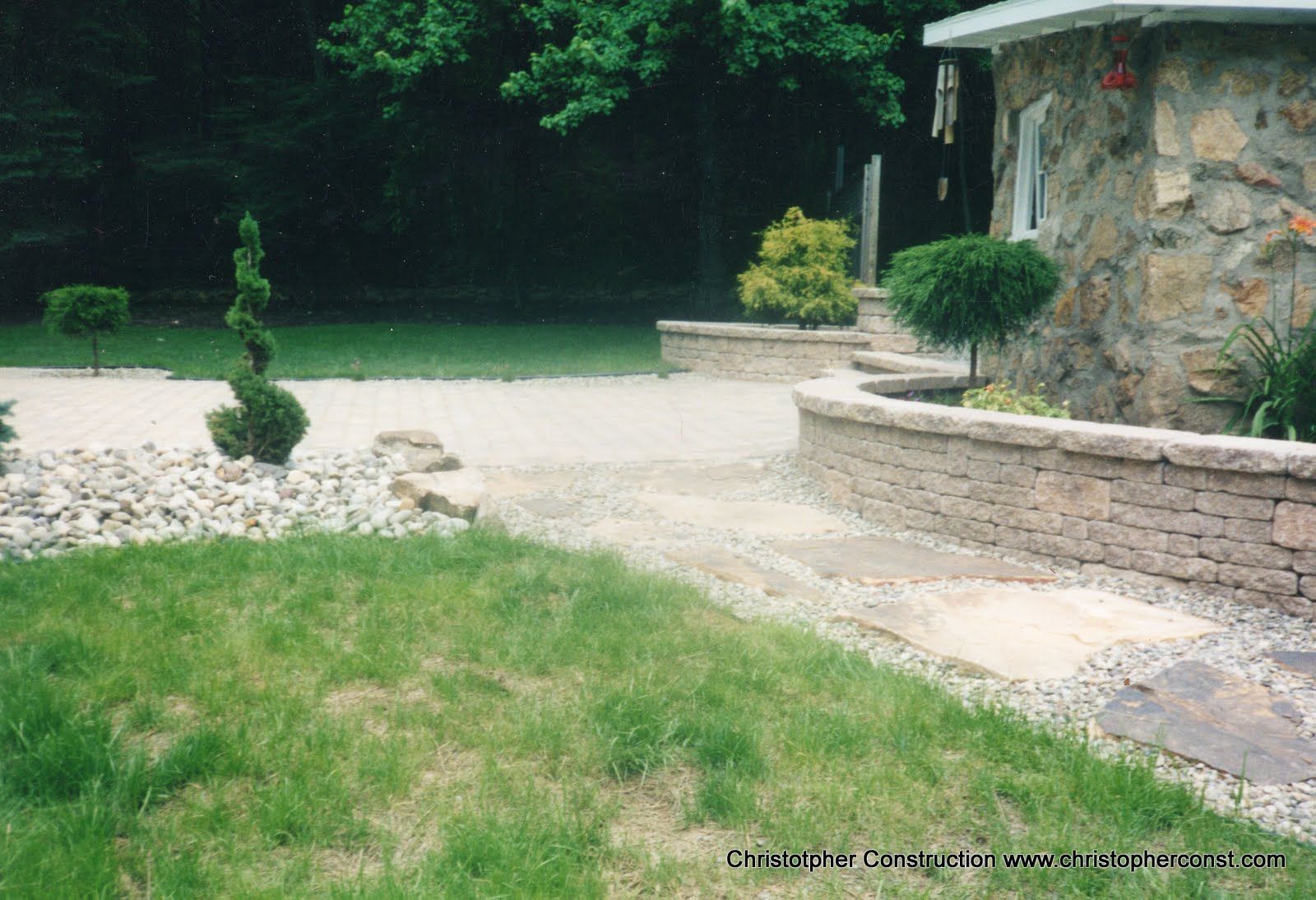 Gravel driveway beside a stone house with manicured shrubs and a green lawn.