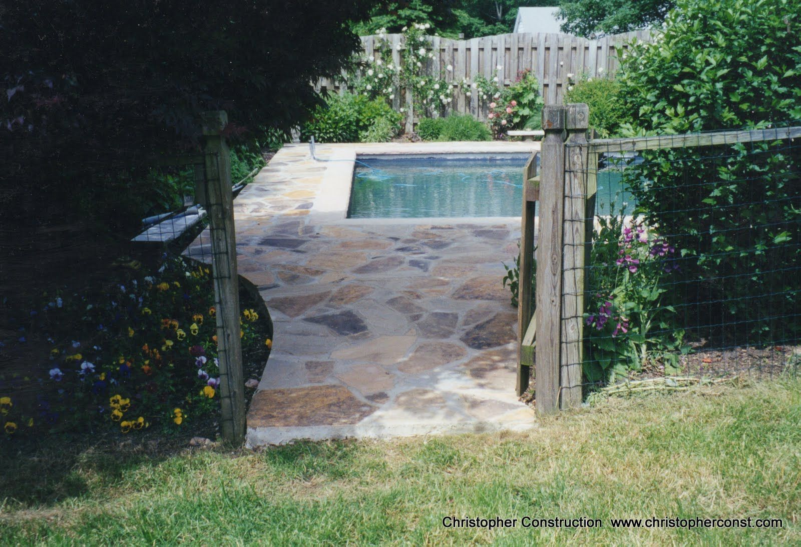 Stone patio with a small rectangular pool, surrounded by greenery and a white fence.