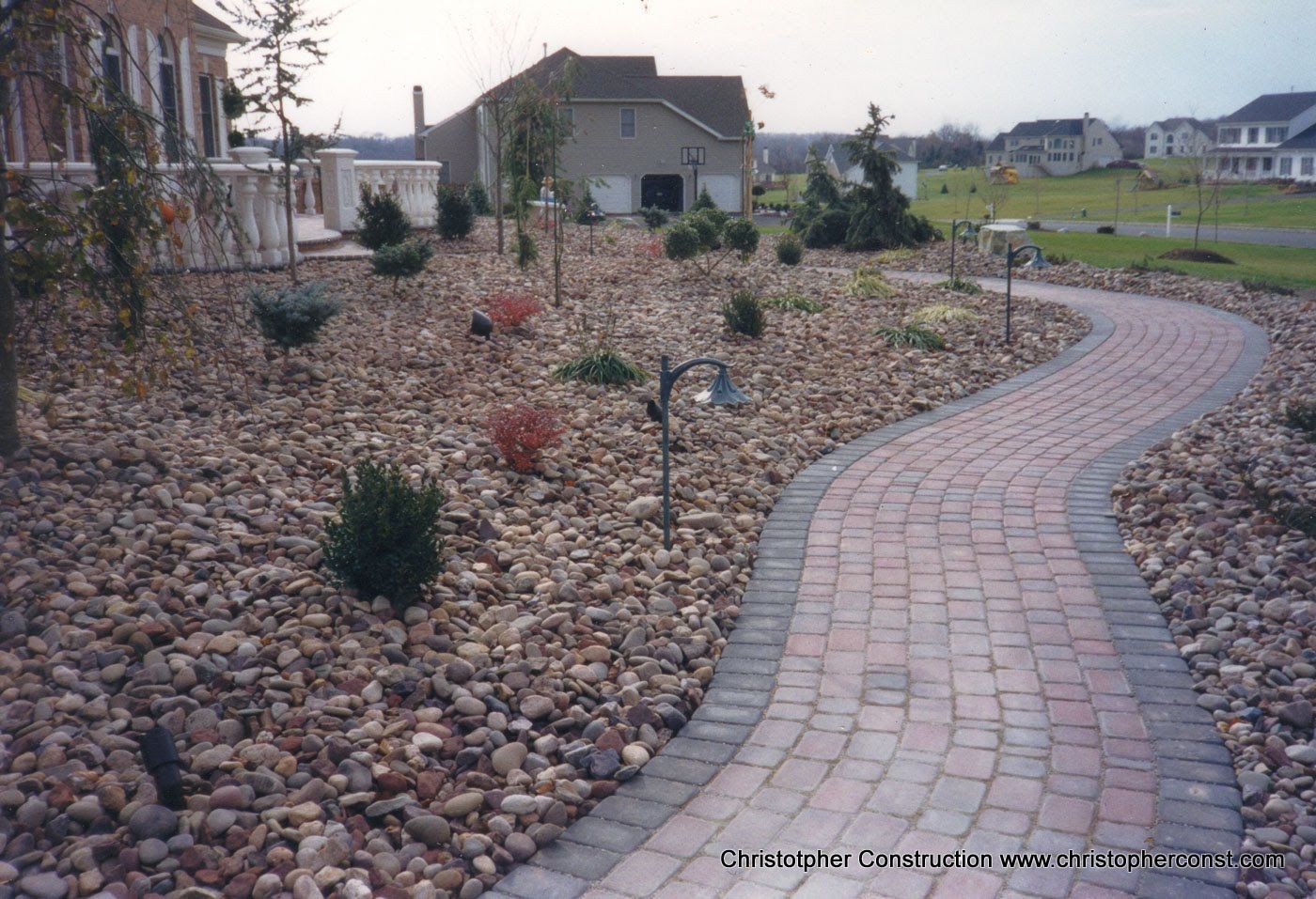 Curved brick walkway through a rock garden with small shrubs and houses in the background
