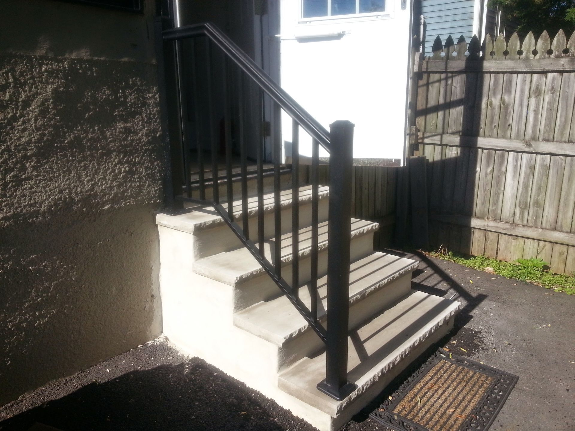 Black metal stairs with railing leading to a white door beside a wooden fence and stucco wall.