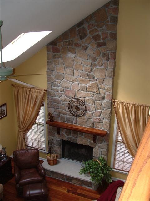 Living room fireplace with stone chimney, leather chair, and curtained windows under a skylight