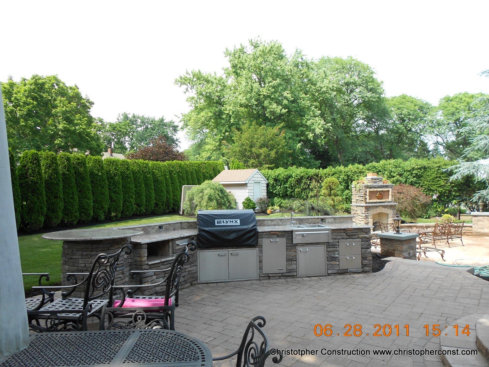 Backyard patio with grill, stone counter, chairs, and lush green hedges and trees.