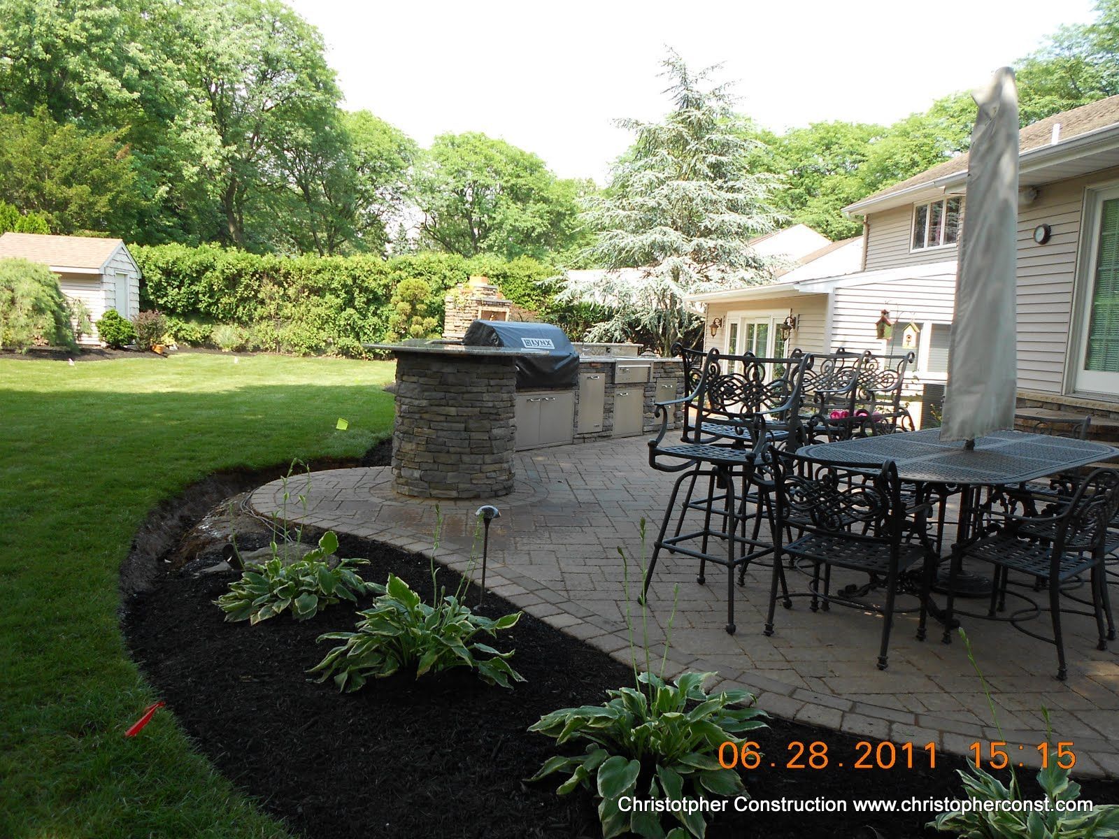 Back patio with black metal table and chairs beside a stone grill and landscaped garden