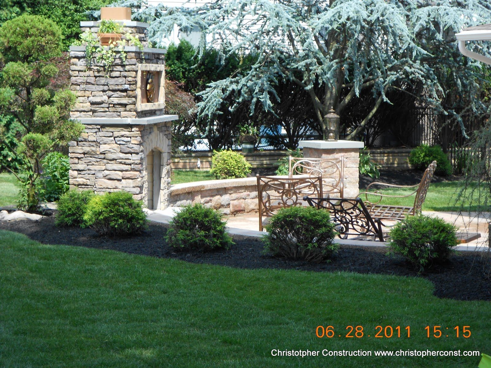 Stone garden gate with pillars, shrubs, and a large tree in a landscaped yard