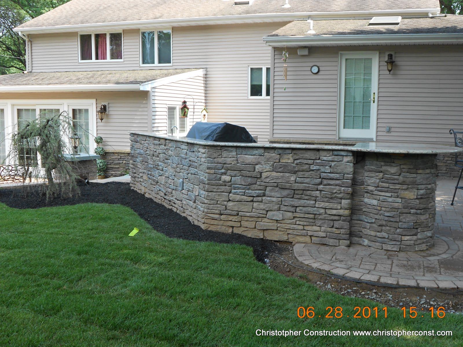 Backyard stone retaining wall and curved patio beside a beige house with a glass door