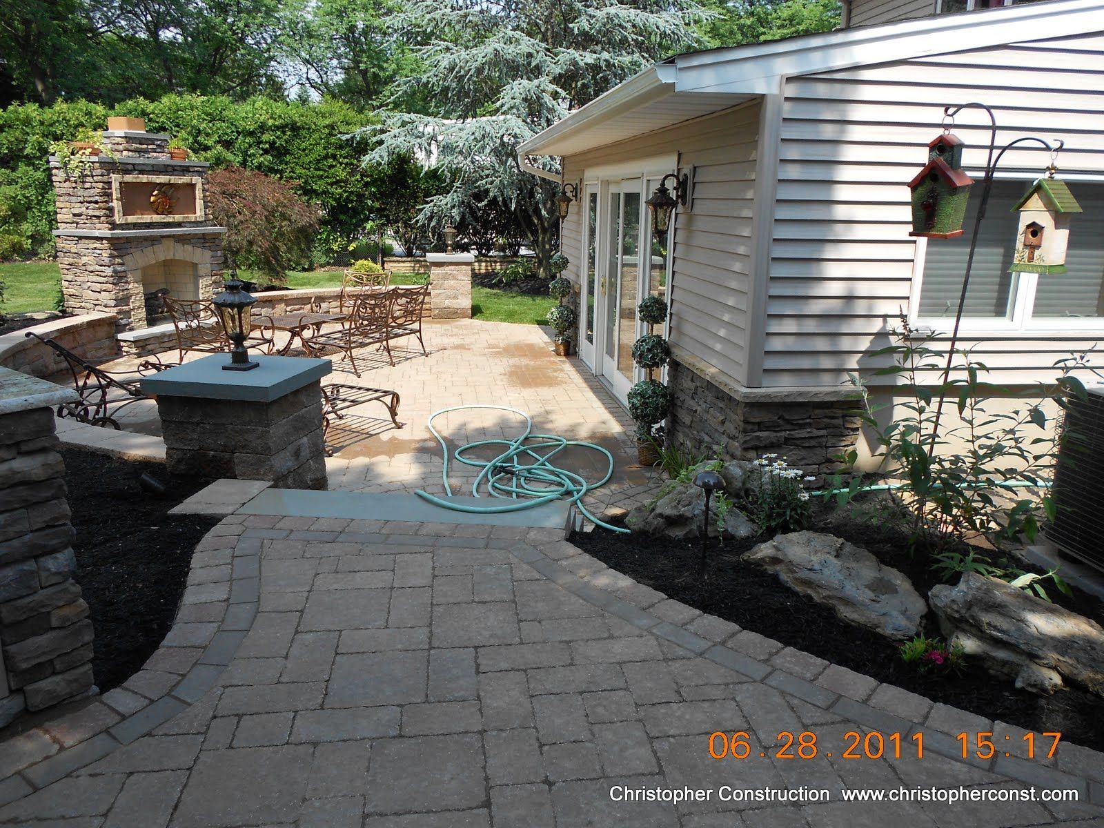 Backyard patio with stone fireplace, paver walkway, and white house with hanging flowers