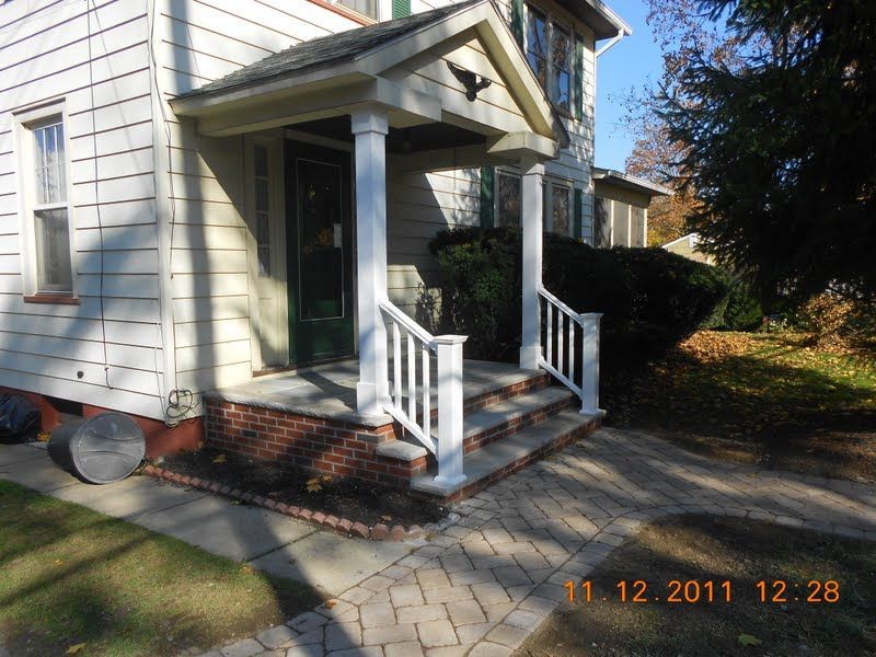 Small front porch with white railings on a beige house, brick steps, and a paver walkway in daylight