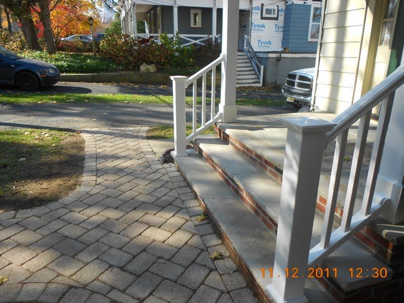 Front steps with white railings leading to a house entrance, beside a sunlit brick walkway and parked cars.