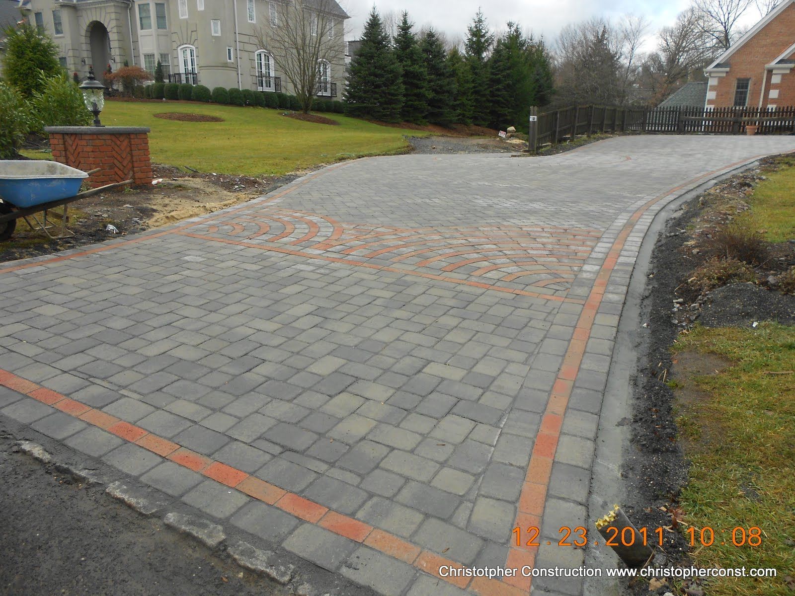 Curved brick-paved driveway beside buildings and grass, with a gray wet patch in the center.