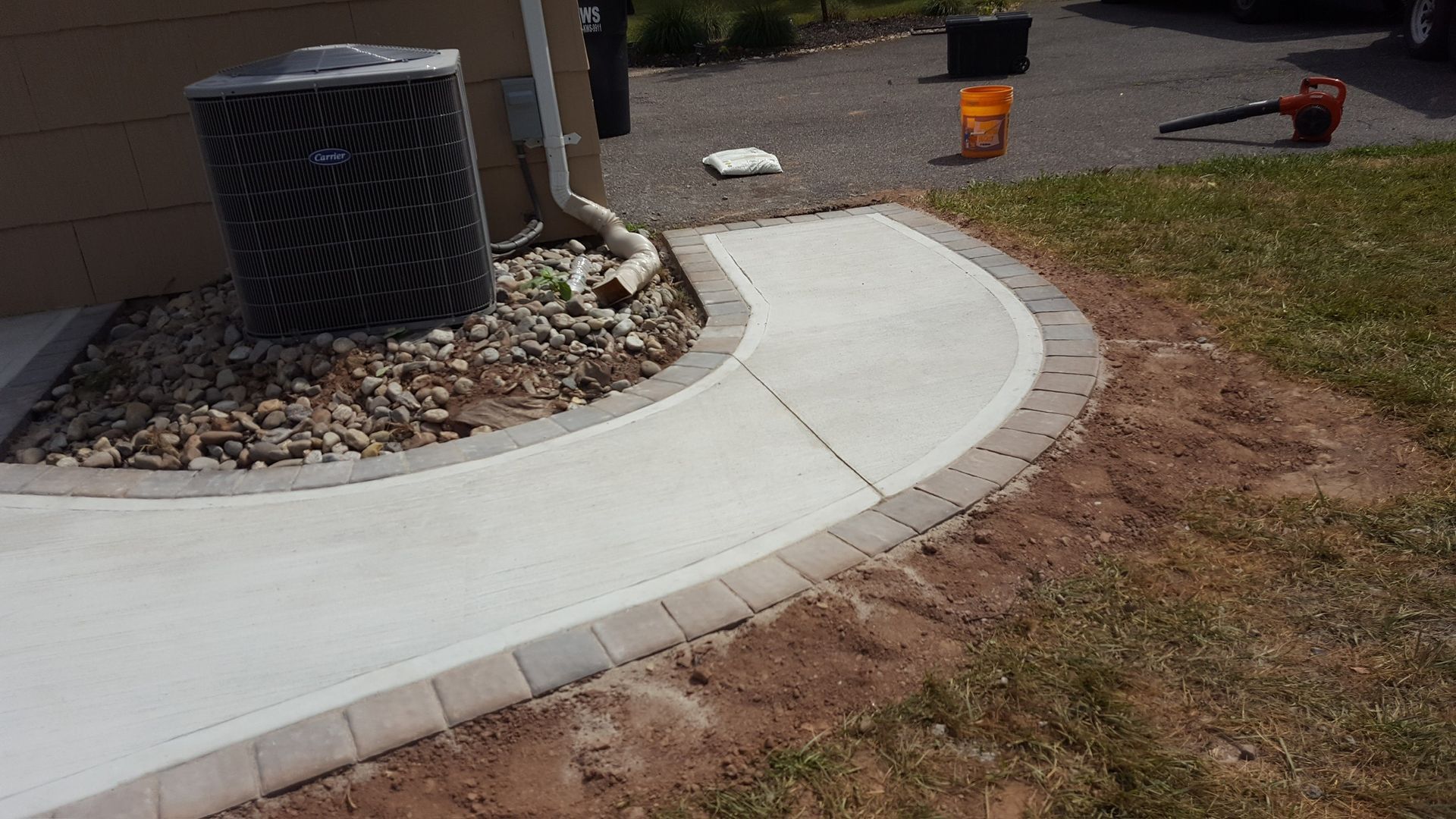 Curved concrete sidewalk beside a house, with gravel landscaping, an AC unit, and a lawn edge.