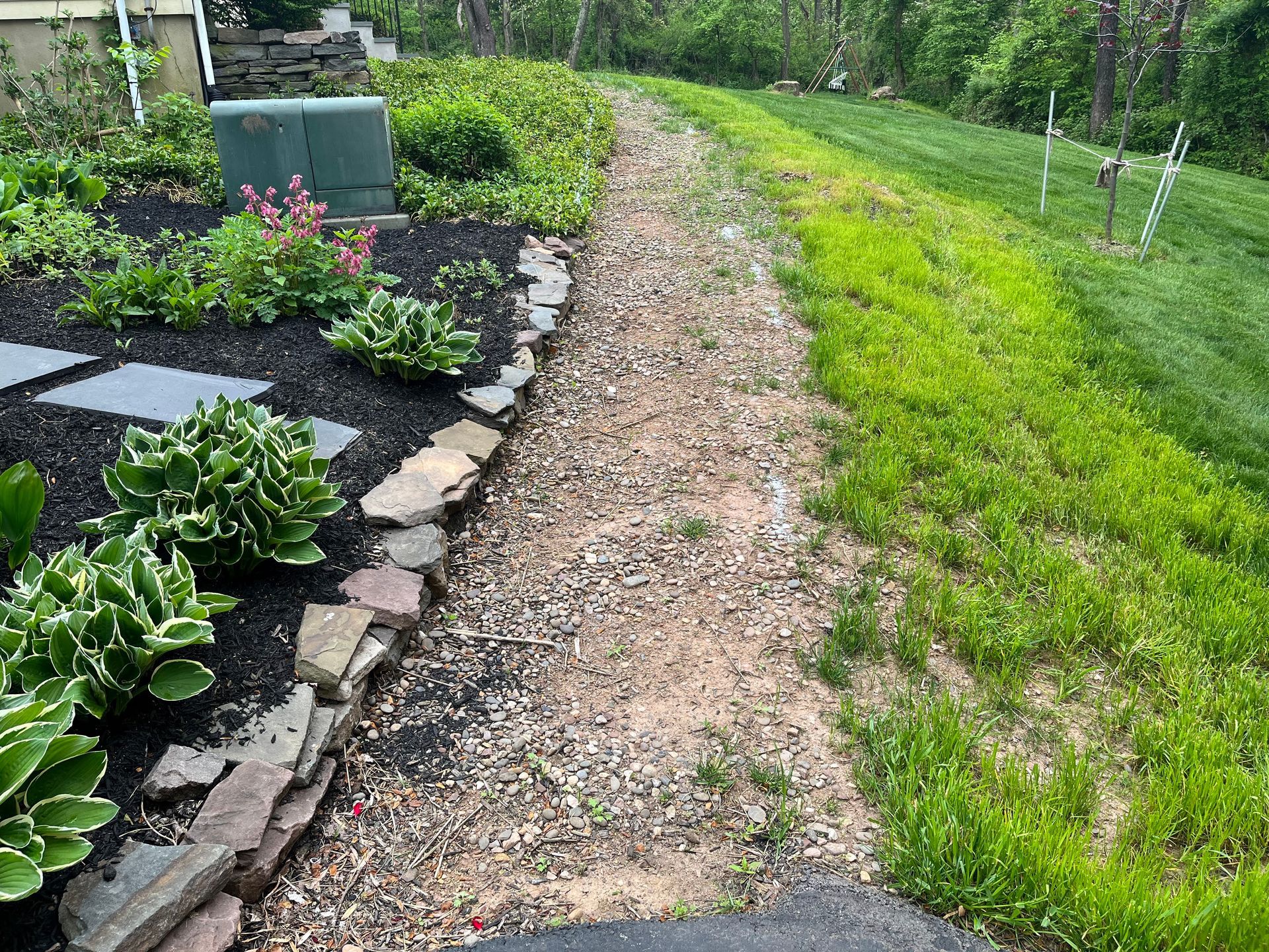 Garden path beside a stone-edged flower bed and green lawn, with shrubs and gravel along a rural fence line
