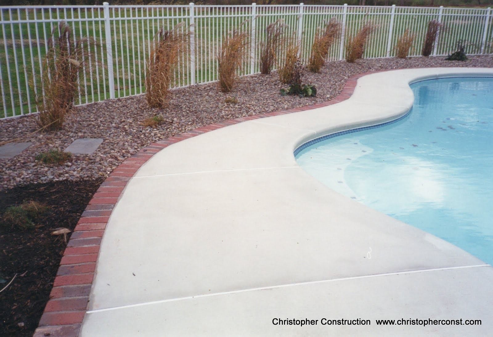 Curved concrete pool deck beside a blue swimming pool, with a brick edge and sparse landscaping along a white fence