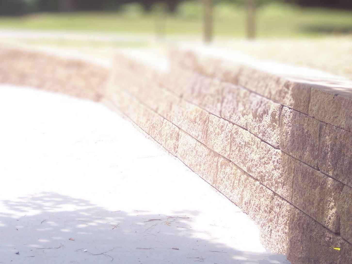 Curved brick retaining wall beside a sunlit paved path with trees in the background