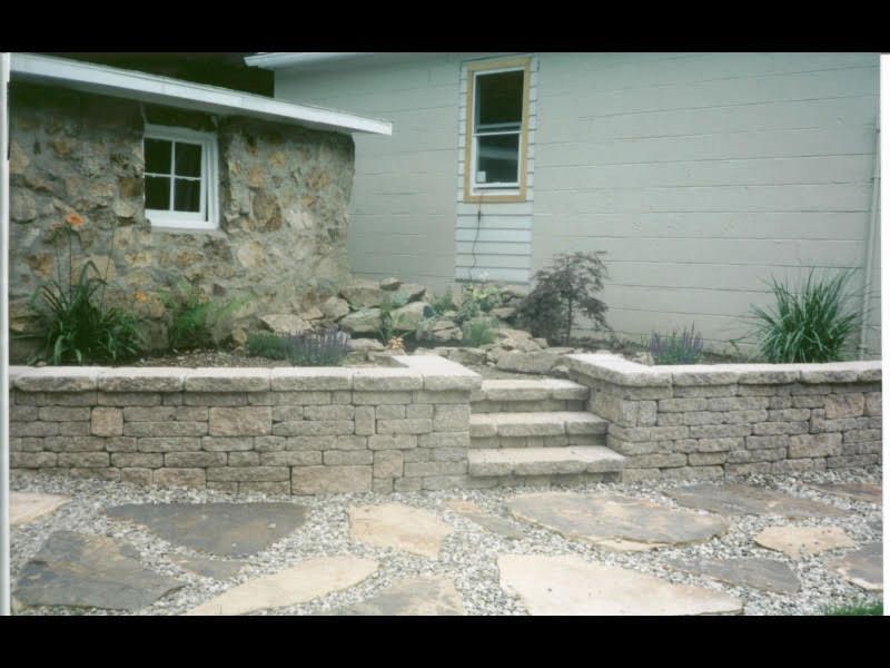 Stone steps and retaining wall leading to a small white house with a stone exterior and garden plants.