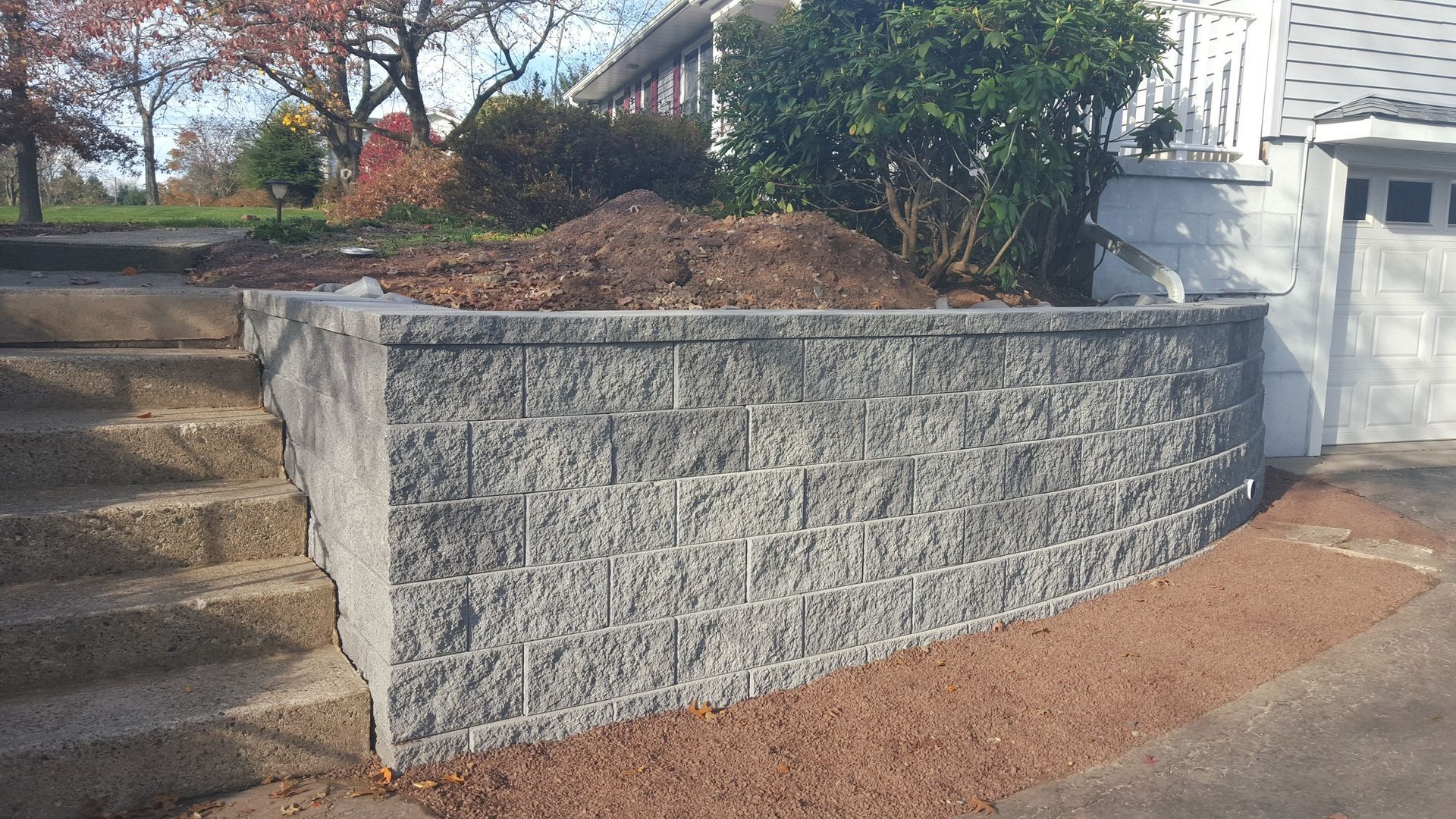Stone retaining wall beside outdoor steps and a white house, with mulch and shrubs in front