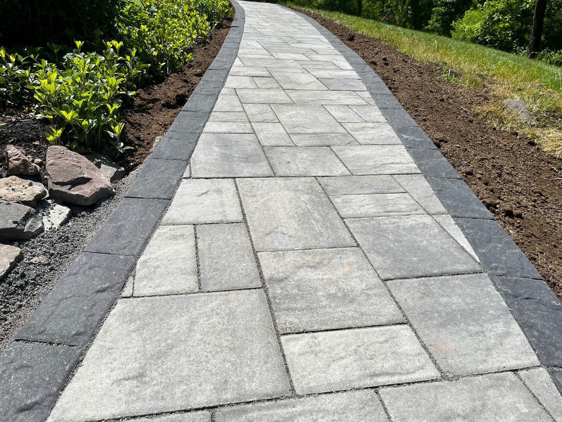 Curved stone walkway bordered by dark edging, grass, and shrubs in a landscaped garden.
