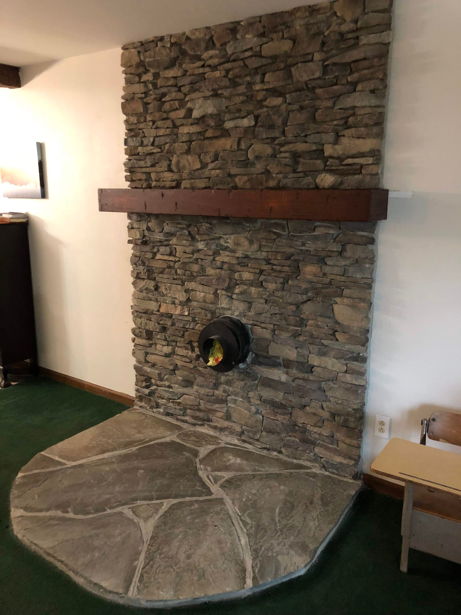 Stone fireplace with a curved hearth and wooden mantel in a room with green carpet.