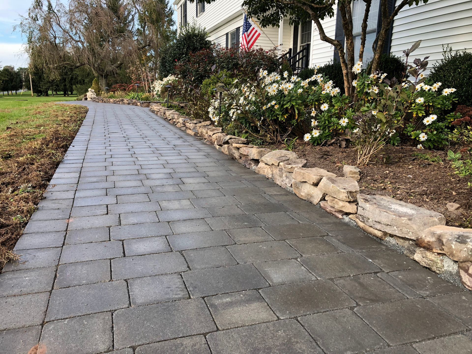 Stone walkway beside a house with flowers, rocks, and an American flag in the yard