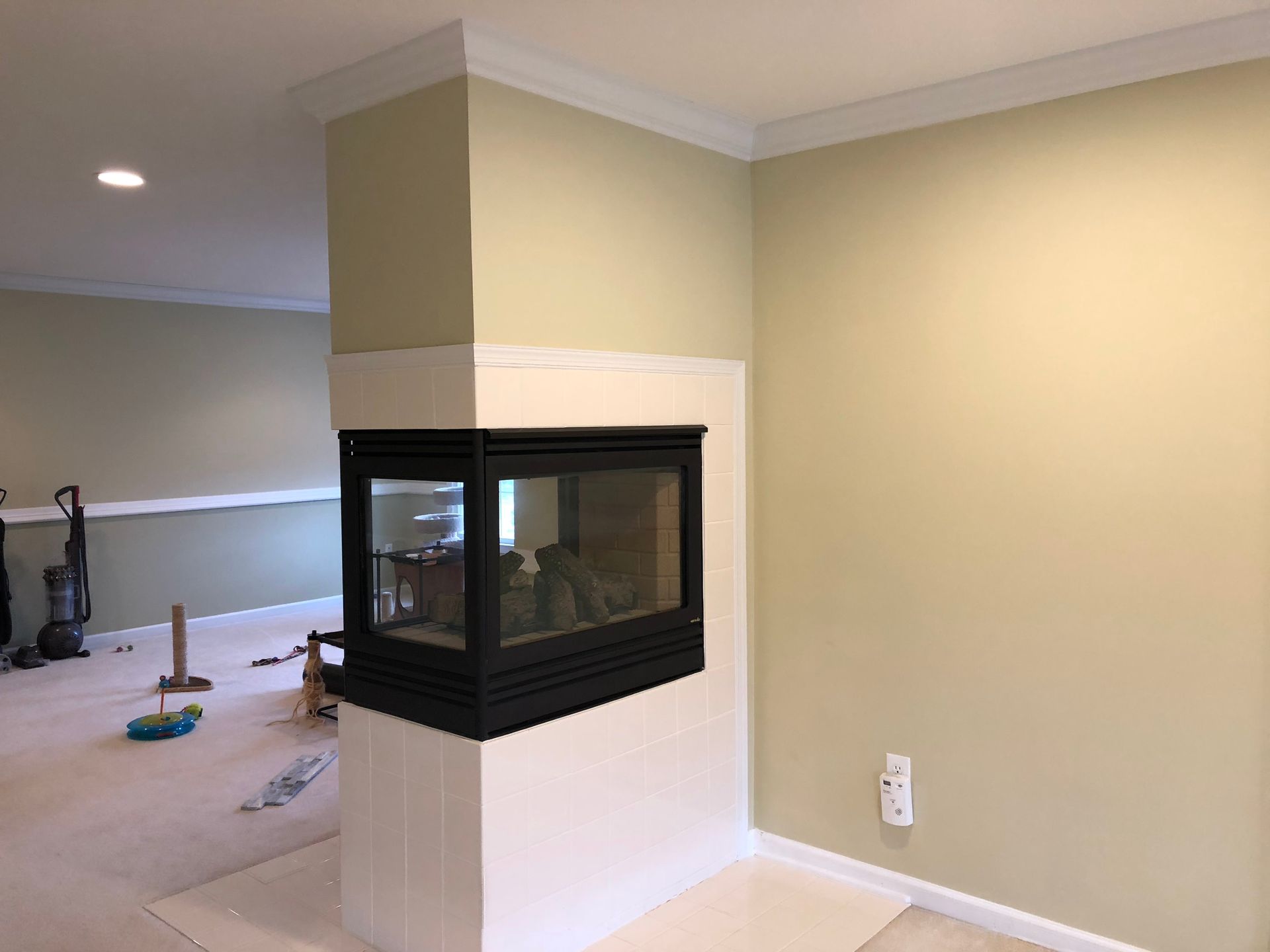 Living room with a corner glass fireplace on a white column and beige walls, opening to a carpeted area.