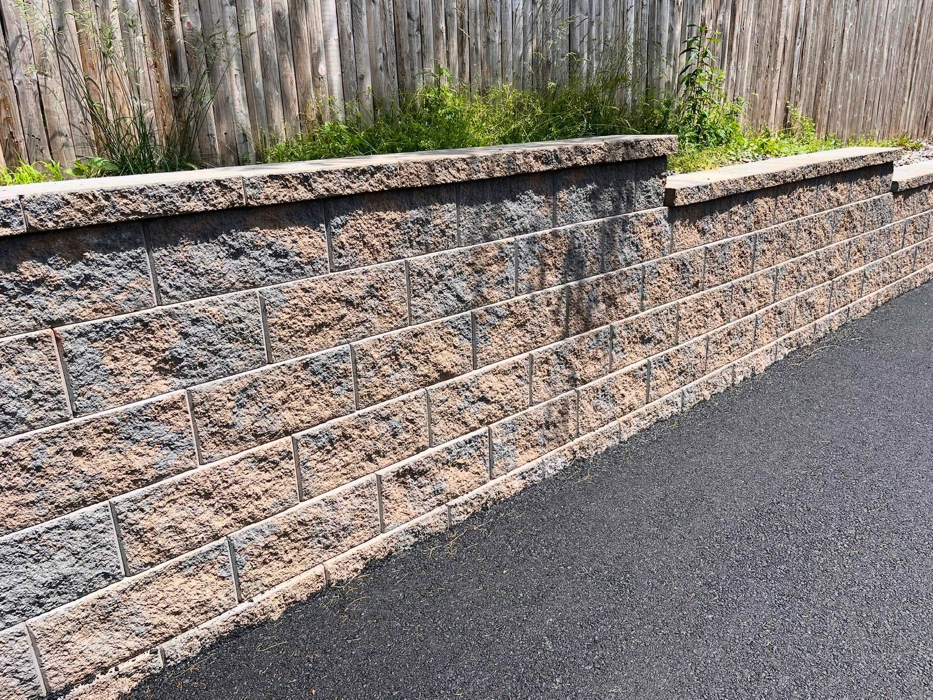 Stone retaining wall beside an asphalt driveway, with a wooden fence and greenery above.