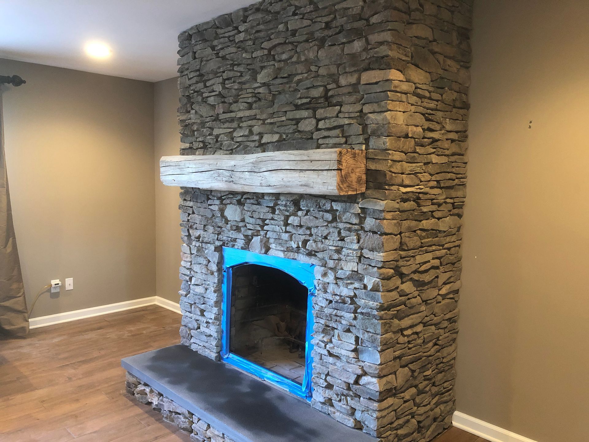 Stone fireplace with a rustic wooden mantel in an empty room with wood floors and beige walls